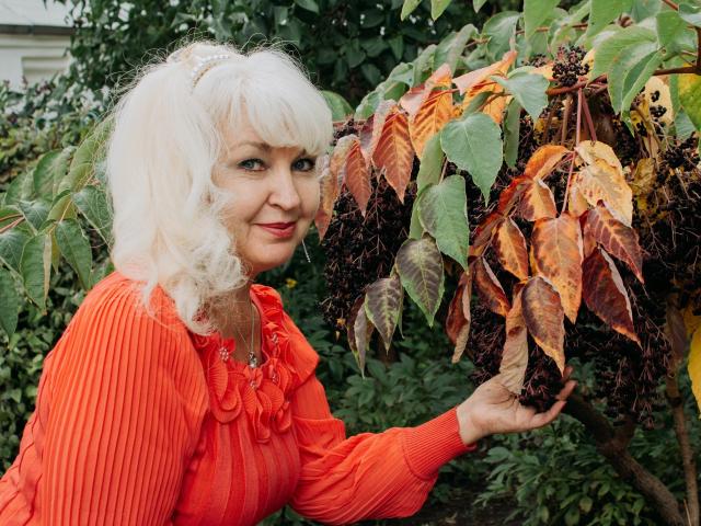 KamaliaSim with curly white hair, wearing an orange ribbed top, holding a leafy tree with brown and yellow leaves, smiling slightly. Her tits are small, she's cute.
