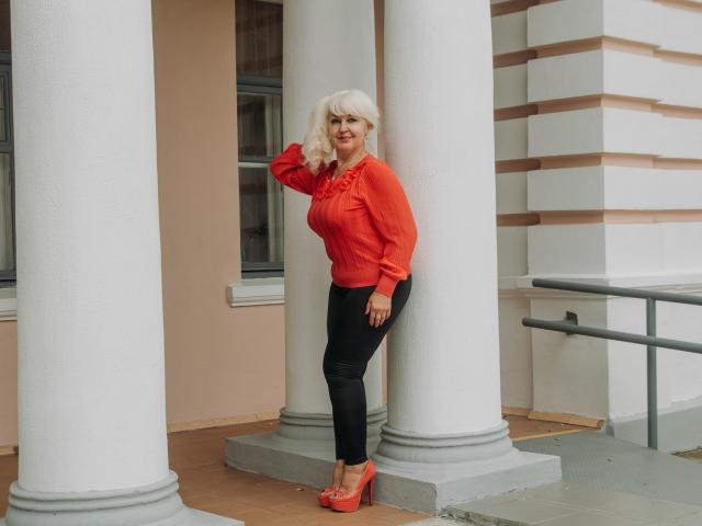 KamaliaSim stands against white columns, wearing a red blouse, black pants, and red heels, with blonde hair, smiling, hand on column. Boobs visible under blouse. Red lipstick.
