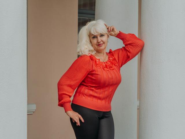 KamaliaSim with white hair, red ribbed shirt, and black pants, standing against a white column, hand on head, smiling, showing her tits subtly.