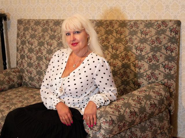 KamaliaSim with white polka-dot blouse, black skirt, and pink lipstick, sitting on floral-patterned sofa, has long blonde hair, pink nails, and a slight smile.