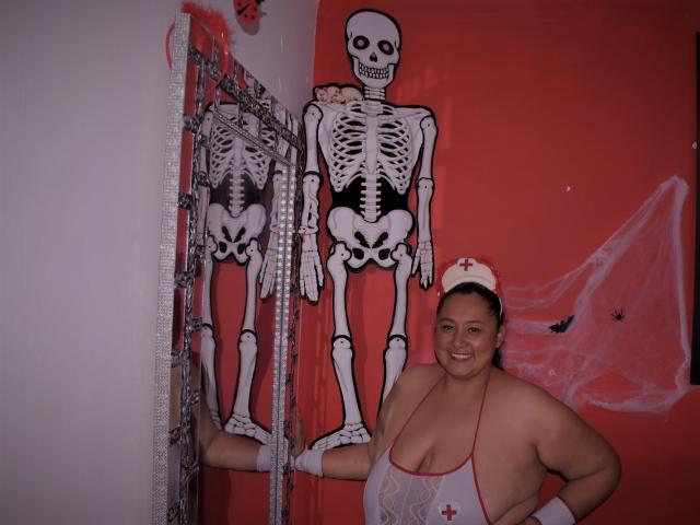 TistMature, con grandi seni, sorridente, che indossa un sexy costume da infermiera, un copricapo bianco e guanti, è in piedi accanto a due decorazioni a forma di scheletro su un muro rosso..