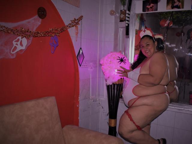 TistMature, obese with large tits and ass, wears a red lace thong and Santa hat, holding a pink star ornament, smiling in a dimly lit room, reflecting in a window.
