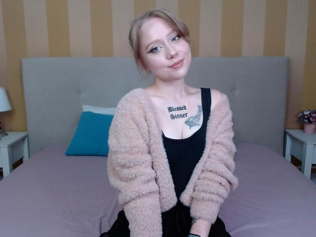 Hayley Trent, com cabelo loiro, vestindo um cardigã rosa felpudo e uma camiseta preta, sentada em uma cama, sorrindo, mostrando seus seios com uma tatuagem acima..