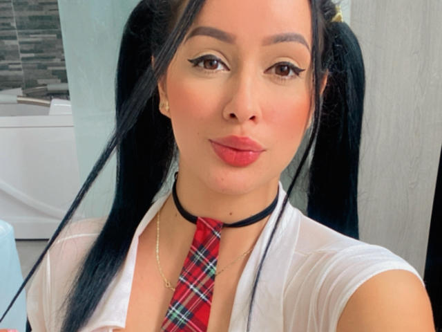 EmilyParkerr has black pigtails, red plaid tie, white shirt, black choker, and red lipstick. She's looking at the camera with winged eyeliner and a slight smile.