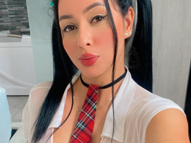 EmilyParkerr has black pigtails, wearing a sheer white shirt with a red plaid tie and black choker, her lips are puckered in a kiss, showing her small tits.