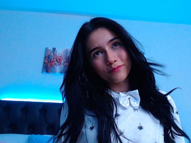 DeisyWest has long black hair, blue eyes, and a small nose with pink lipstick. She's wearing a white shirt with black star patterns, in a blue-lit room.