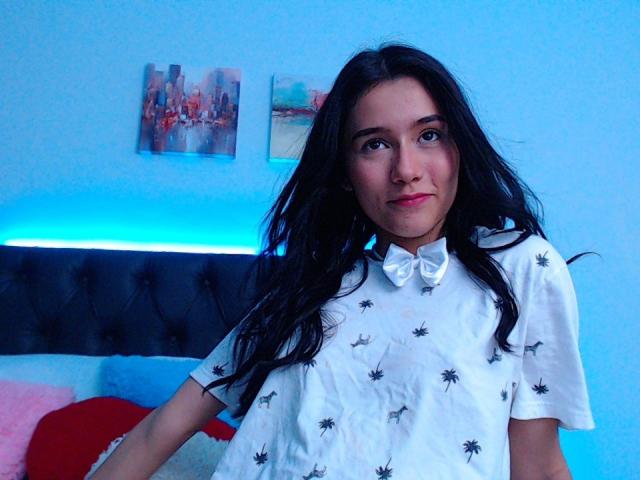 DeisyWest with long black hair, wearing a white shirt with black spider patterns, sitting in a blue-lit room, smiling softly, blue eyes, light skin, medium build.