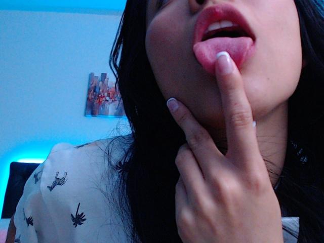 DeisyWest's face with her finger in her pink, glossy lips, dark hair, blue lighting, white shirt with black bird patterns, nail polish, slightly open mouth.