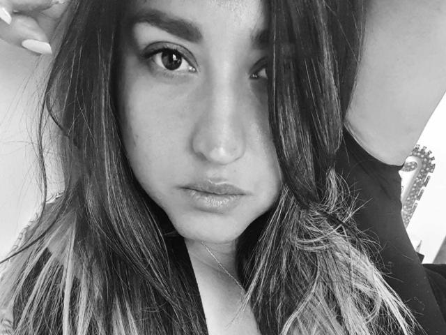 El rostro de NinaZorya, una fotografía en blanco y negro, con cabello largo y ondulado, cejas oscuras, labios brillantes y una expresión seria. Está levantando su brazo derecho, mostrando sus uñas largas..