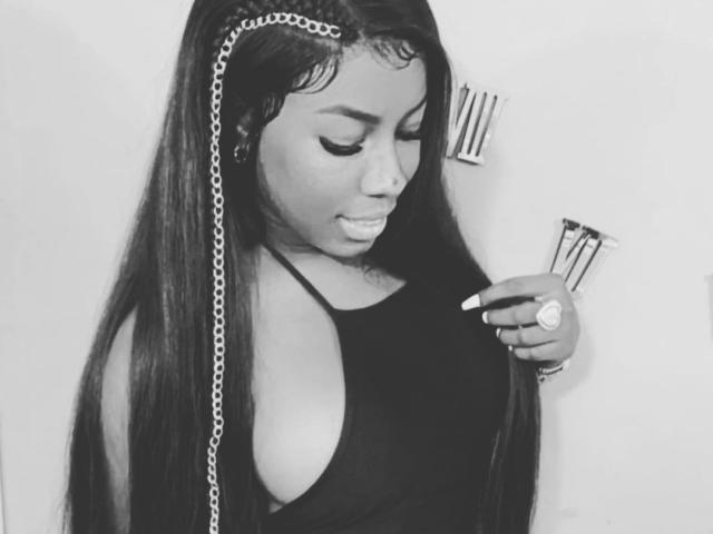 KatyBomb, avec de longs cheveux lisses, portant un débardeur noir, a une chaîne dans ses cheveux, regarde vers le bas, tient une bague, et ses seins sont légèrement visibles. Photo en noir et blanc..