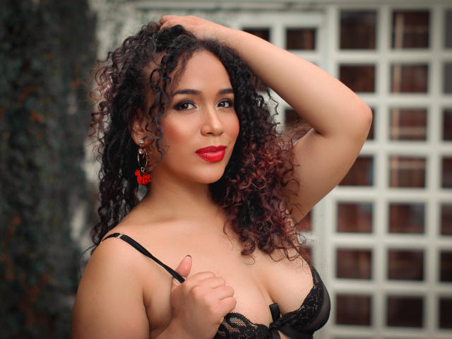 SelynRobert, con cabello negro rizado, lápiz labial rojo y un sujetador de encaje negro, con la mano en el cabello, mostrando sus pechos, aretes rojos y una sonrisa segura..