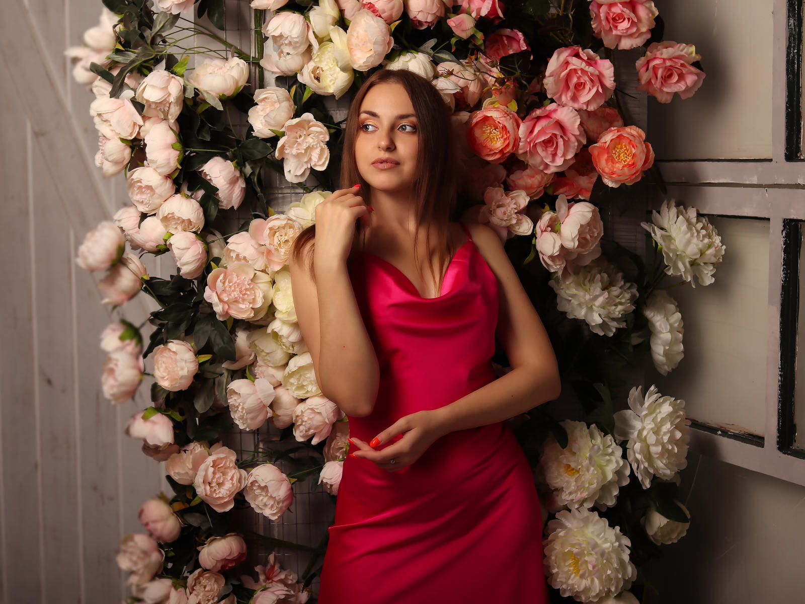 Jessica, avec des cheveux roux, portant une robe rose, debout devant un mur de fleurs roses et blanches, touchant son menton, regardant vers le haut, avec sa main droite posée sur son ventre..
