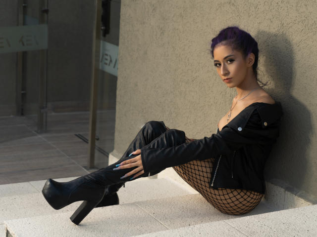 JuliaJobs with purple hair, sitting against a wall, wearing a black jacket, fishnet pants, and black boots, showing her tits, with a confident, sexy expression.