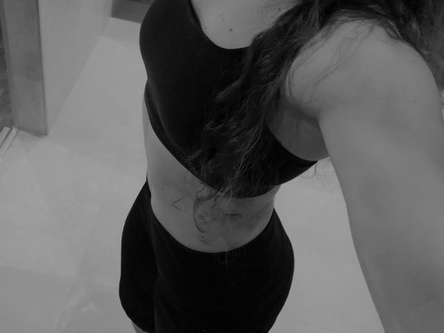 WishForMe met golvend donker haar, gekleed in een zwarte sportbeha en zwarte broek, met een tatoeage zichtbaar op haar onderbuik, op een zwart-wit foto..