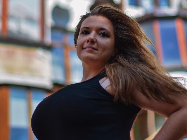 PatriciaSyde with long brown hair, wearing a black tank top, showing her big tits, looking down with a slight smile, her hair blowing in the wind.