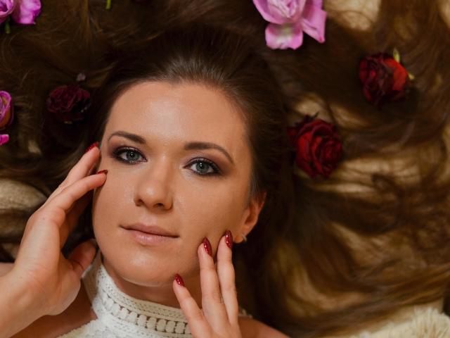Het gezicht van PatriciaSyde, met blauwe ogen, bruin haar en rode nagellak, liggend op een bed van bloemen, met handen die haar wangen aanraken, en een witte gebreide kraag dragend..