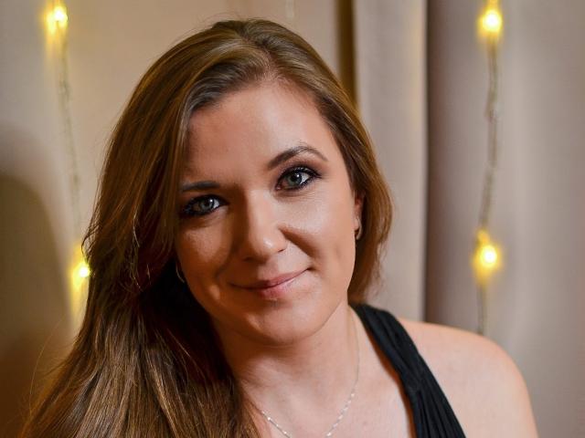 PatriciaSyde, con i capelli castani, gli occhi verdi e un sorriso discreto, indossa una maglia nera e guarda direttamente l'obiettivo. Il suo seno non è visibile, ma il suo viso sì..