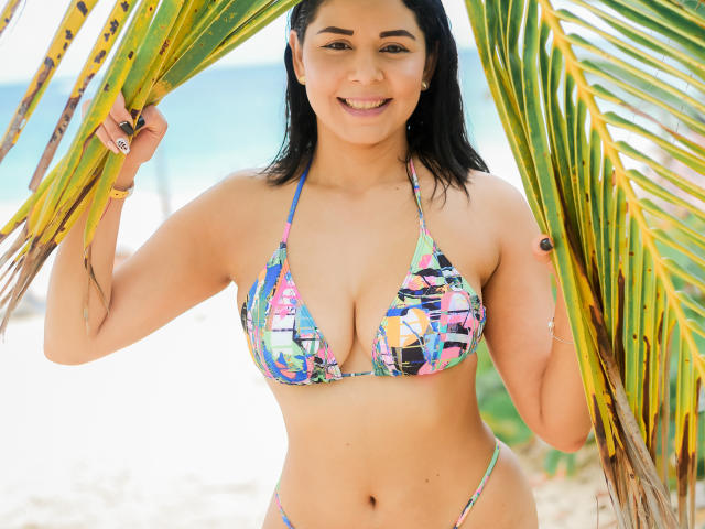 EvaMonn, com seios médios, vestindo um biquíni colorido e estampado, segurando folhas de palmeira, sorrindo, tem pele bronzeada, cabelo preto e um corpo curvilíneo e sexy, em pé em uma praia ensolarada..
