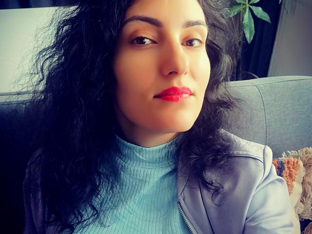 AddictedToUTS has curly black hair, red lipstick, and a light blue turtleneck, wearing a gray jacket. She's sitting on a gray couch with a plant in the background.