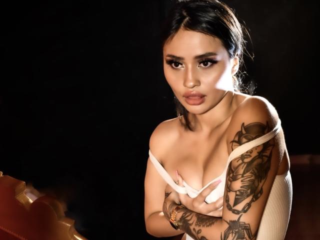 AmbarRiians, com pele morena, cabelos escuros e tatuagens, puxa uma blusa branca para baixo, expondo seus seios, olhando sedutivamente para a câmera. Ela tem uma expressão de lábios franzidos..