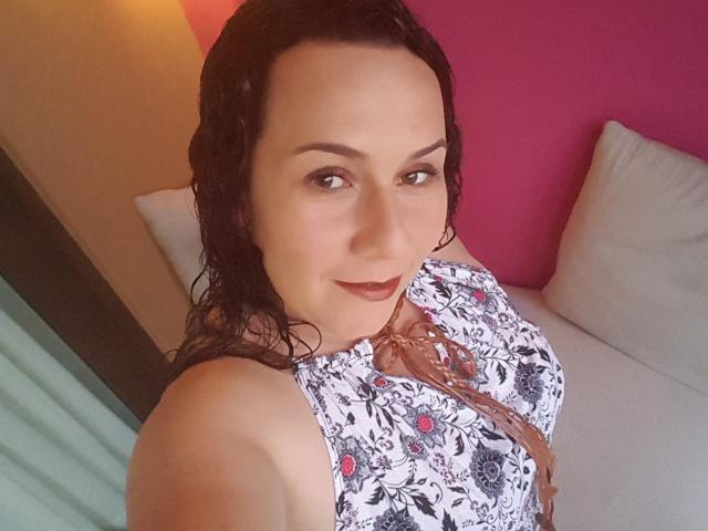 SamySexxiy mit nassen, schwarzen Haaren, trägt ein Blumenkleid mit einem rosa Schleifchen, lächelt in die Kamera und zeigt ihre Brüste. Im Hintergrund ist eine rosa Wand zu sehen..