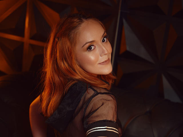 MollyMarmalade with red hair, cute smile, and sexy gaze, wearing a sheer black top, looking over her shoulder, showing her shiny ass and small tits. Warm, dark, and moody.