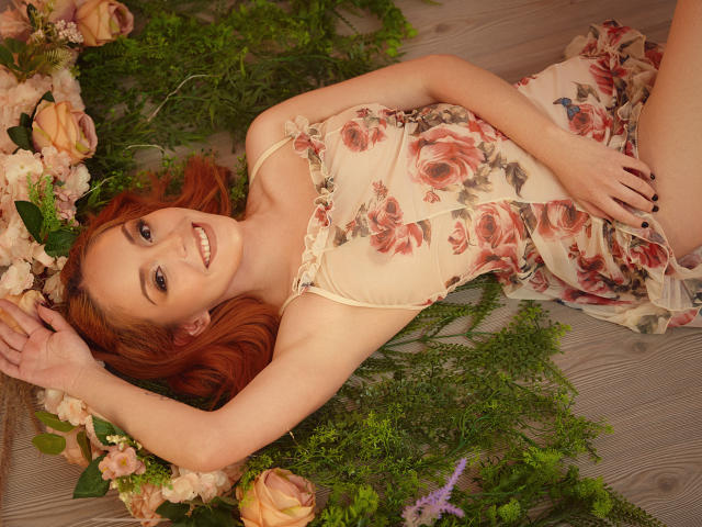 MollyMarmalade with red hair, smiling, lying on floral arrangements, wearing a floral dress, hands above her head, on wooden floor, looking up, cute, pretty, tits slightly visible.