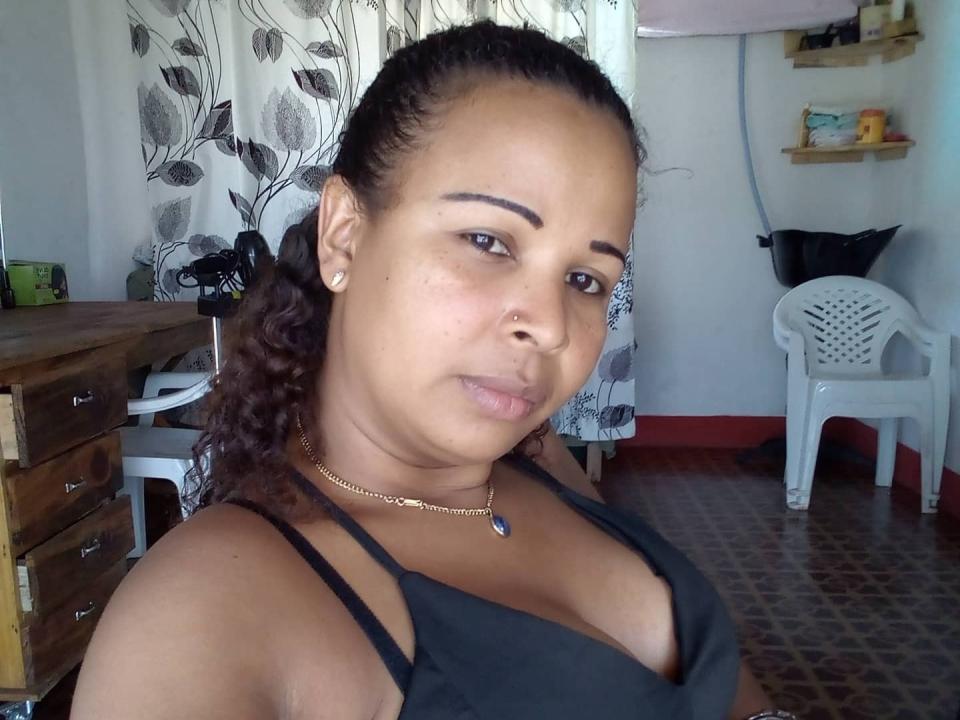 HannaSofis, plus size, com cabelo escuro, sorrindo, vestindo uma blusa de malha laranja neon que expõe seus seios grandes e uma tanga preta, sentada em uma cama rosa..