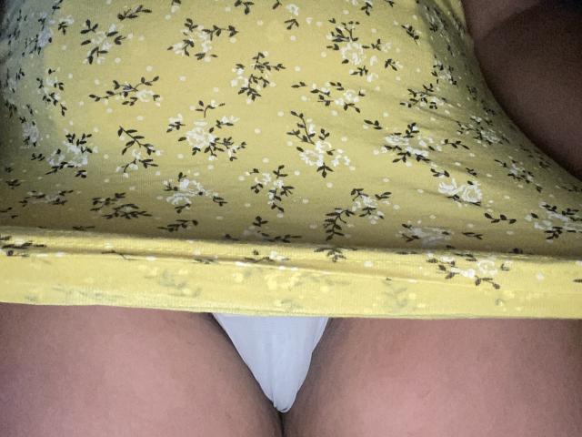 La robe fleurie jaune de DearLola est soulevée, révélant sa culotte blanche et ses cuisses légèrement écartées. Sa robe est ornée de petites fleurs noires et blanches, et elle a une peau claire..