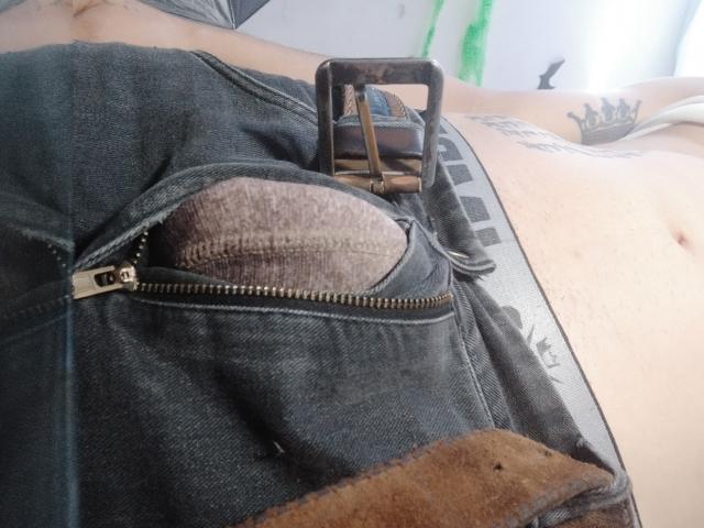 Jean déchiré d'LatinAndres, laissant apparaître un panneau marron, texturé et sans entrejambe. Boucle de ceinture en métal, tatouages sur le côté et le bas du ventre, peau blanche et ventre plat..