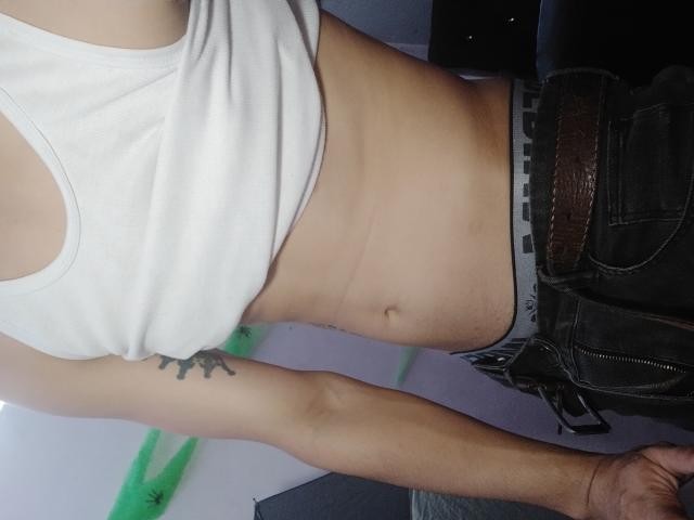 A camiseta branca de LatinAndres, que mostra um abdômen fino e plano com um umbigo pequeno, e uma tatuagem no braço esquerdo. Ele está vestindo calças escuras com um cinto marrom..