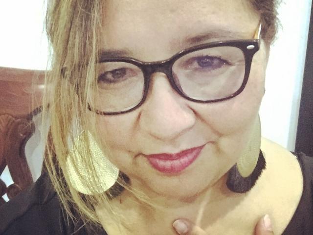 AmbarSimmons has glasses, blonde hair, and is wearing large gold earrings with black trim. She's smiling with pink lipstick, showing a slight double chin and black top.