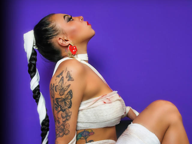 NenaBlack with braided ponytail, red earrings, white bandage wrap, tattoos on arm, and red lipstick, leaning back against purple background, showing her ass and tits.