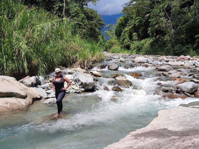 YiyaHot69 stands in a rocky stream, wearing a black wetsuit and a white sunhat, with her foot on a large rock, water swirling around her.