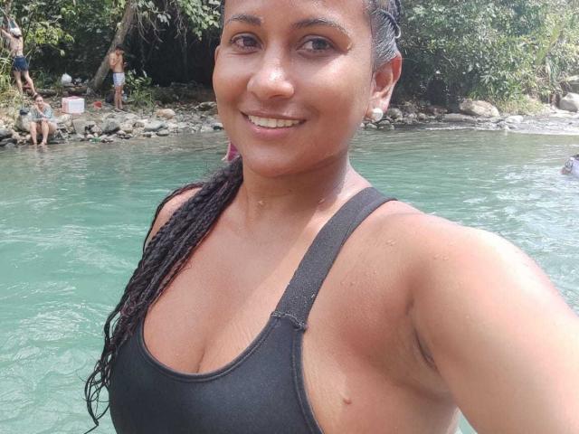 YiyaHot69 with wet brown skin, black tank top, and long braided hair, showing her tits and smiling at the camera in a green, rocky river.