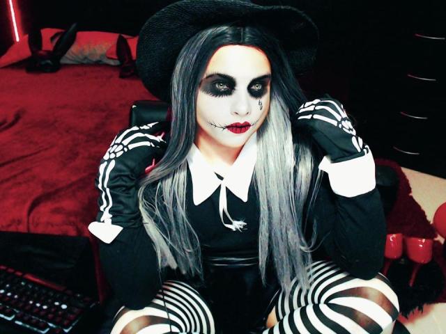 CamilaFetishDirty con il viso truccato di un pallido bianco, occhiaie scure, rossetto rosso, cappello da strega nero, lunghi capelli grigi, pantaloni a righe nere e bianche e guanti neri, seduta sul letto..