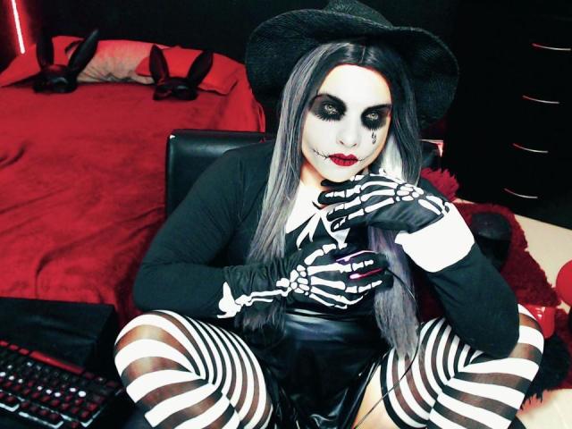 CamilaFetishDirty, avec un maquillage pâle, un chapeau noir et des gants rayés, assise sur un lit, portant un haut noir et un legging rayé, avec un nœud papillon noir sur sa poitrine..