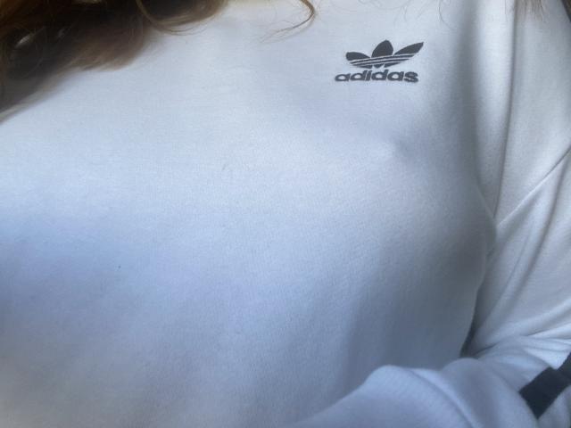 Rubbi's white Adidas shirt shows a small black Adidas logo on the upper chest, with a slight visible nipple. Her brown curly hair is partially visible.