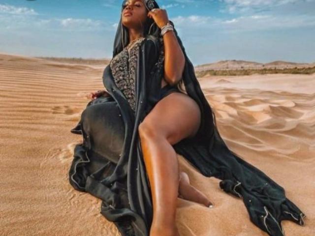 KatyBomb, peau foncée, longs cheveux noirs, portant une robe noire à motifs, assise sur du sable dans le désert, montrant ses fesses, les jambes croisées, tenant un téléphone noir, ciel bleu au-dessus..
