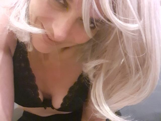 Natachatte with blonde hair, looking down, has her tits in a black lace bra, slightly leaning forward, her face partially obscured by her hair.