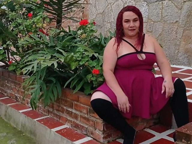 SamySexxiy, una donna corpulenta con i capelli rossi e grandi seni, è seduta su un davanzale di mattoni, indossa un abito magenta e calze nere fino al ginocchio, e sorride alla telecamera..
