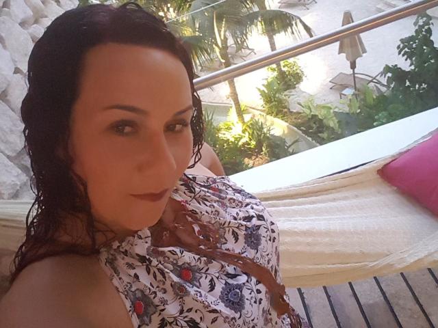 SamySexxiy, con i capelli scuri e umidi, indossa un abito floreale, mostrando il décolleté, seduta in un'amaca bianca, che guarda la fotocamera con un leggero sorriso..