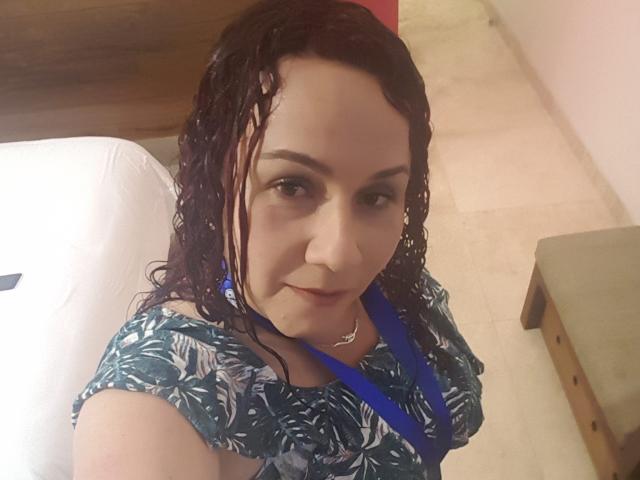 SamySexxiy, con i capelli castani bagnati, indossa una maglietta blu a motivi, una collana blu e un piercing al naso, e guarda la fotocamera con un leggero sorriso. Il suo seno non è visibile..