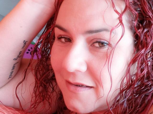 SamySexxiy, com cabelo ruivo encaracolado e molhado, pele clara e uma tatuagem no braço direito, olhando para a câmera com um leve sorriso, mostrando seu pequeno nariz e ombros nus..