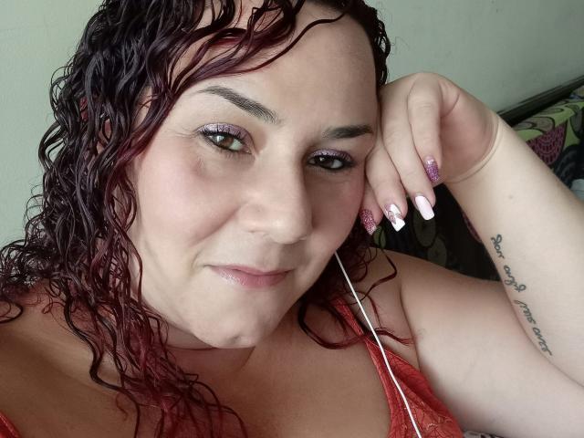 SamySexxiy mit nassen, lockigen roten Haaren, rosa glitzernden Augenlidern und rosa glitzernden Nägeln, blickt in die Kamera, leichtes Lächeln, rotes Spitzenoberteil, Tattoo am rechten Unterarm, Faden im Mund..