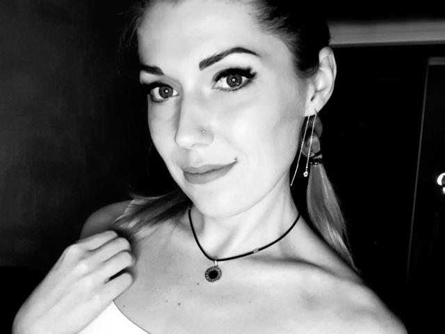 NikiSport, con un sorriso malizioso, si tiene una spalla, ha un piercing al naso, eyeliner scuro e una collana con una gemma. Indossa un top senza spalline e orecchini lunghi..