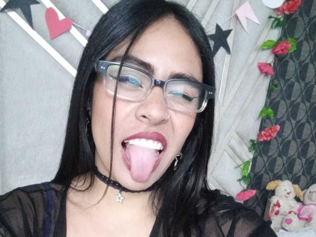 StellaBeckker ha i capelli neri, porta gli occhiali e un piercing al naso. Ha la lingua fuori e gli occhi chiusi, indossa una collana nera e una maglia trasparente che mostra il seno..