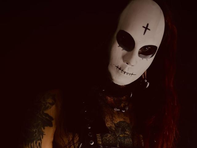 EscariotNEeva met een wit masker, donkere ooggaten, een genaaide mond, een kruis op het voorhoofd, rode dreadlocks, een zwarte kanten top en tatoeages, in gedimd licht..