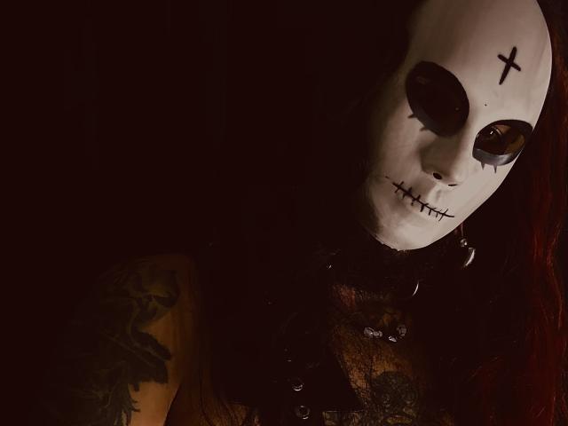 EscariotNEeva, con una maschera bianca a forma di teschio, fori oculari scuri, una croce nera sulla fronte, labbra cucite, un tatuaggio sul braccio sinistro e capelli rosso scuro, indossa pizzo nero..