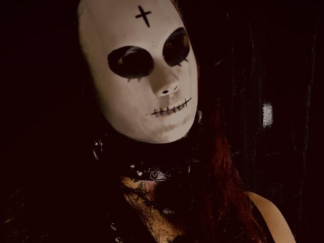 EscariotNEeva con una maschera bianca inquietante, occhi neri, bocca cucita e una croce sulla fronte, indossa pizzo nero e trecce rosse, su uno sfondo scuro..